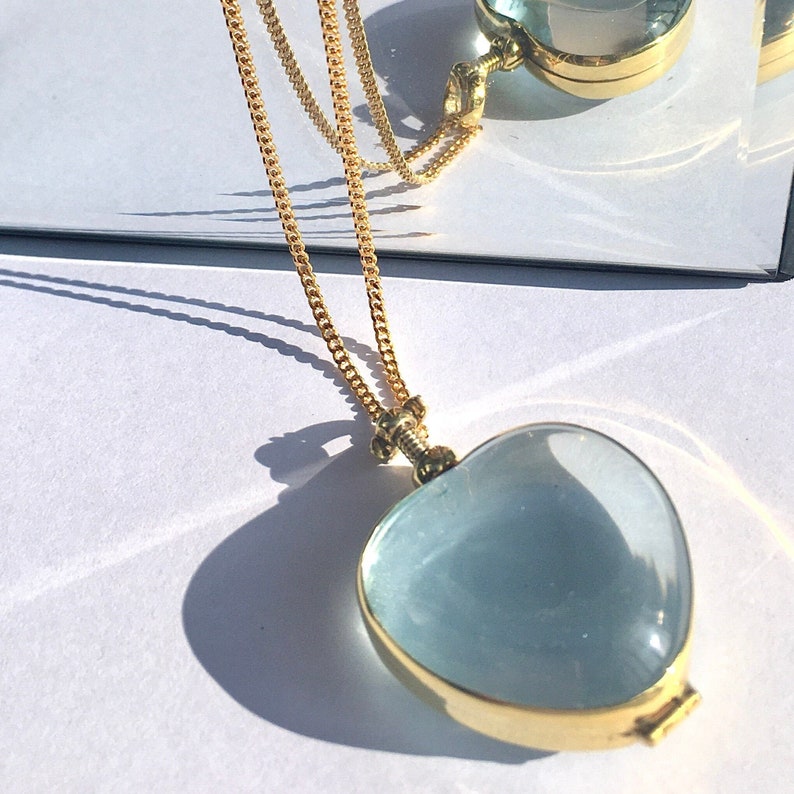 May include: A gold chain necklace with a heart-shaped locket. The locket is made of clear glass and has a gold frame. The locket is open and the inside is visible.