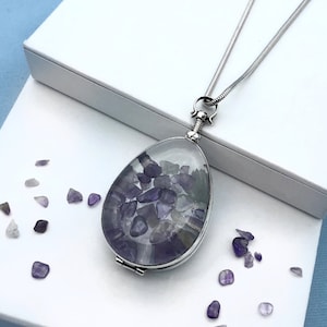May include: A silver chain necklace with a teardrop-shaped locket. The locket is open and filled with small purple and clear crystals.
