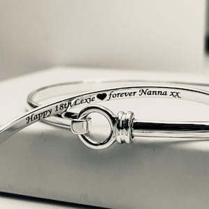 May include: Two silver bangles are shown in close-up. One is inscribed with "Happy 18th Lexie" and "forever Nanna xx" in black lettering. The other bangle is partially visible. The bangles are on a white surface.