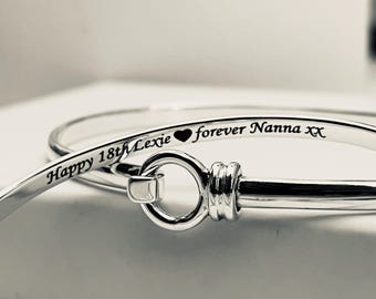 May include: Close-up of two silver-colored bangles. One bangle has the inscription "Happy 18th Lexie" and "forever Nanna xx" engraved in black. The other bangle is partially visible. The bangles are on a white surface.