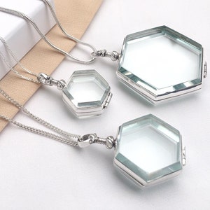May include: Three silver-toned hexagonal pendant necklaces. Each pendant has a clear glass front and back, with a hinged opening. The necklaces are displayed on a white surface with a light tan fabric.