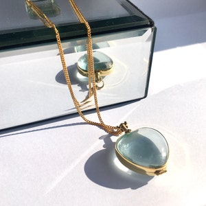 May include: A gold chain necklace with a heart-shaped locket pendant. The locket is made of clear glass and has a light blue tint.