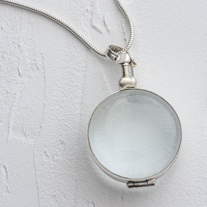 Op de afbeelding: Een zilverkleurige vergrootglas hanger ketting met een heldere, ronde lens. De hanger heeft een scharnierend ontwerp en hangt aan een delicate slangenketting. Het vergrootglas heeft een diameter van ongeveer 5 cm.