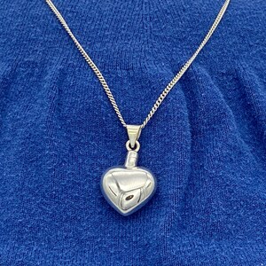 Puede incluir: Un collar de plata con un colgante en forma de coraz&oacute;n en una cadena de plata. El colgante es liso y brillante.