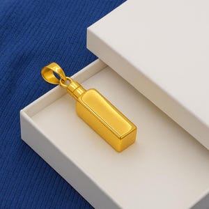 May include: A gold-colored pendant in the shape of a rectangular bottle, displayed in an open white box. The pendant has a loop at the top for a chain. The background is a textured blue surface.