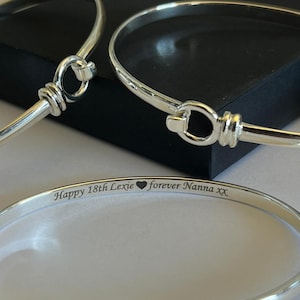 May include: Three silver bangles with a hook and eye clasp are shown. One bangle has the inscription "Happy 18th Lexie ❤️ forever Nanna XX". The bangles are displayed on a black surface.