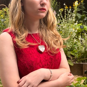 May include: A woman wearing a red lace dress with a gold heart-shaped locket necklace. The locket has a clear glass front and is filled with small white flowers.