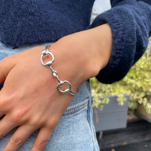 May include: A silver bangle bracelet with a snaffle bit design. The bracelet is on a person's wrist.