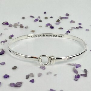 Puede incluir: Un brazalete de plata con la inscripción "Love you to the moon and back!". El brazalete tiene una superficie texturizada y un cierre de gancho. El fondo es blanco, con piedras decorativas moradas y grises dispersas.