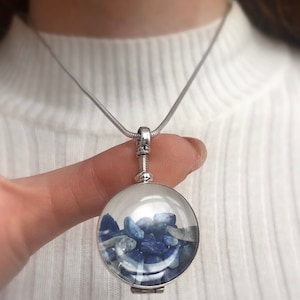 Op de afbeelding: Een zilverkleurige ketting met een ronde, transparante hanger gevuld met blauwe stenen. De hanger hangt aan een dunne, zilveren ketting. De ketting wordt gepresenteerd tegen een witte achtergrond.