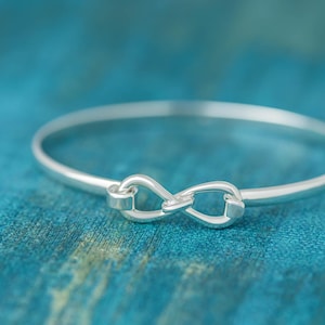 Puede incluir: Un brazalete de plata con un dije en forma de símbolo de infinito. El brazalete está sobre un fondo azul.