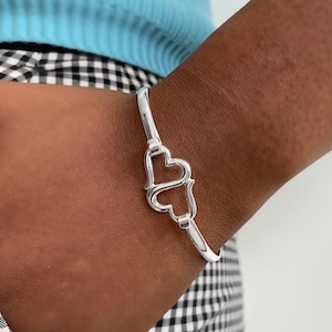 Könnte beinhalten: Ein silberfarbenes Armband mit herzförmigem Verschluss. Das Armband mit dem Design aus ineinander verschlungenen Herzen wird am Handgelenk getragen. Der Hintergrund zeigt ein hellblaues Oberteil und ein schwarz-weißes Karomuster.