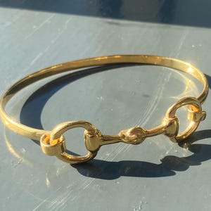 May include: A gold-toned bangle bracelet with a horse bit design. The bracelet is simple and elegant.