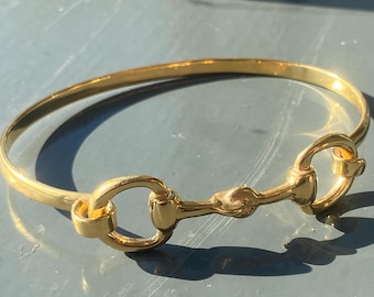 May include: A gold-toned bangle bracelet featuring a horse bit design. The bracelet is simple and elegant.