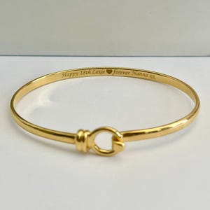 May include: A gold-coloured bangle bracelet with the engraved text "Happy 18th Lexie forever Nanna XX". The bracelet has a clasp closure and a smooth, polished finish. The jewellery is a simple, elegant design.