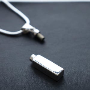Puede incluir: Un colgante rectangular plateado con una pequeña tapa en un extremo, sobre una superficie negra. En el fondo se ve una cadena plateada, conectada a una pieza plateada similar. El colgante tiene una superficie pulida y reflectante.
