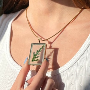 May include: A gold chain necklace with a rectangular pendant featuring a pressed green leaf inside.