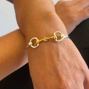 May include: A silver bangle bracelet with a gold-toned horse bit charm. The bracelet is open and has a simple design.