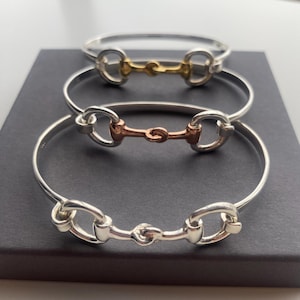 May include: Three silver bangle bracelets with a horse bit design. The top bracelet has a gold bit, the middle bracelet has a rose gold bit, and the bottom bracelet has a silver bit.