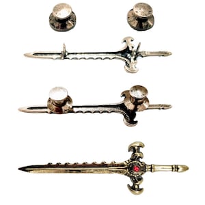 May include: A collection of antique-style sword pins and cufflinks. The pins are silver-toned with a detailed sword design, and the cufflinks feature a similar sword design with a red jewel. The set includes two round-topped cufflinks.