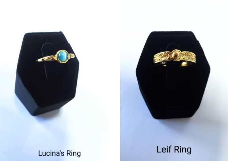 May include: Two gold rings displayed on black velvet stands. The first ring, "Lucina's Ring," features a gold band with a light blue gemstone. The second ring, "Leif Ring," is a gold band with an ornate design and a small, round, orange gemstone.