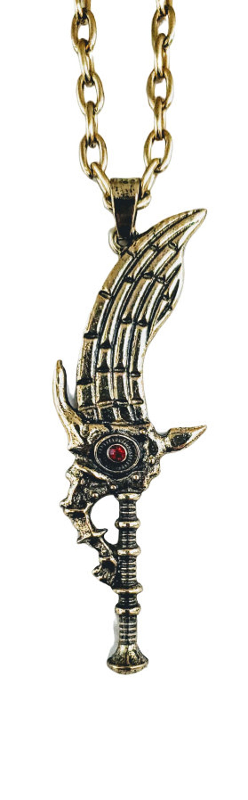 May include: A gold-colored pendant necklace featuring a stylized sword design. The pendant has a wing-like shape with intricate detailing, a red gem, and a chain. The necklace is a fantasy-themed accessory.