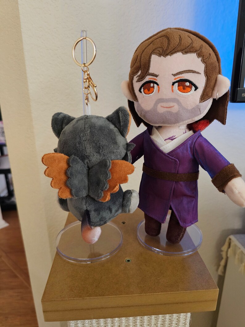 May include: A plush toy keychain featuring a grey and orange winged creature with a gold chain and ring. The keychain is displayed next to a plush toy of a man with brown hair, a purple robe, and a beard. Both toys are on clear plastic display stands.