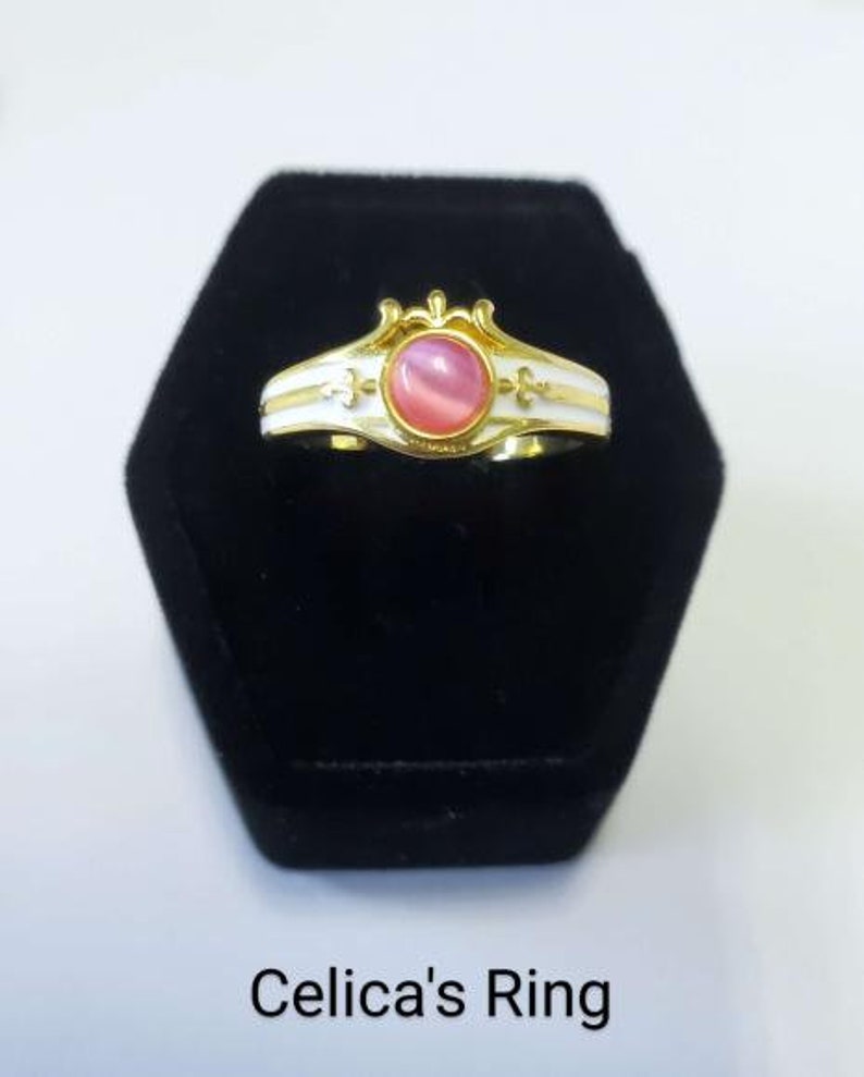 May include: A gold-colored ring with a pink gemstone center, white enamel accents, and decorative flourishes. The ring is displayed on a black velvet surface. The text "Celica's Ring" is at the bottom.