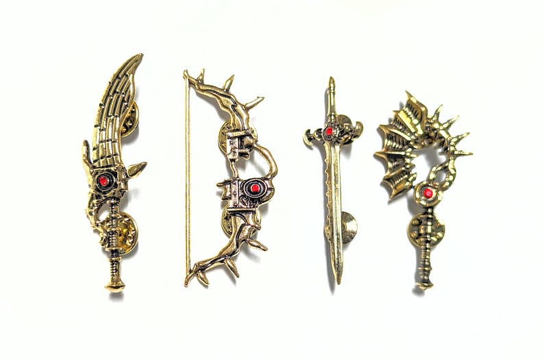 May include: Four gold-colored metal pins in fantasy weapon designs. The pins include a sword, a bow and arrow, a key, and a blade-like weapon. Each pin has a red jewel accent.