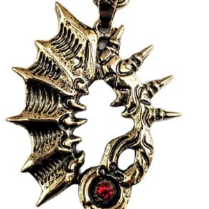 May include: A bronze-colored pendant necklace featuring a stylized key design. The pendant has a detailed, ornate pattern with a red gemstone and is suspended from a matching chain.
