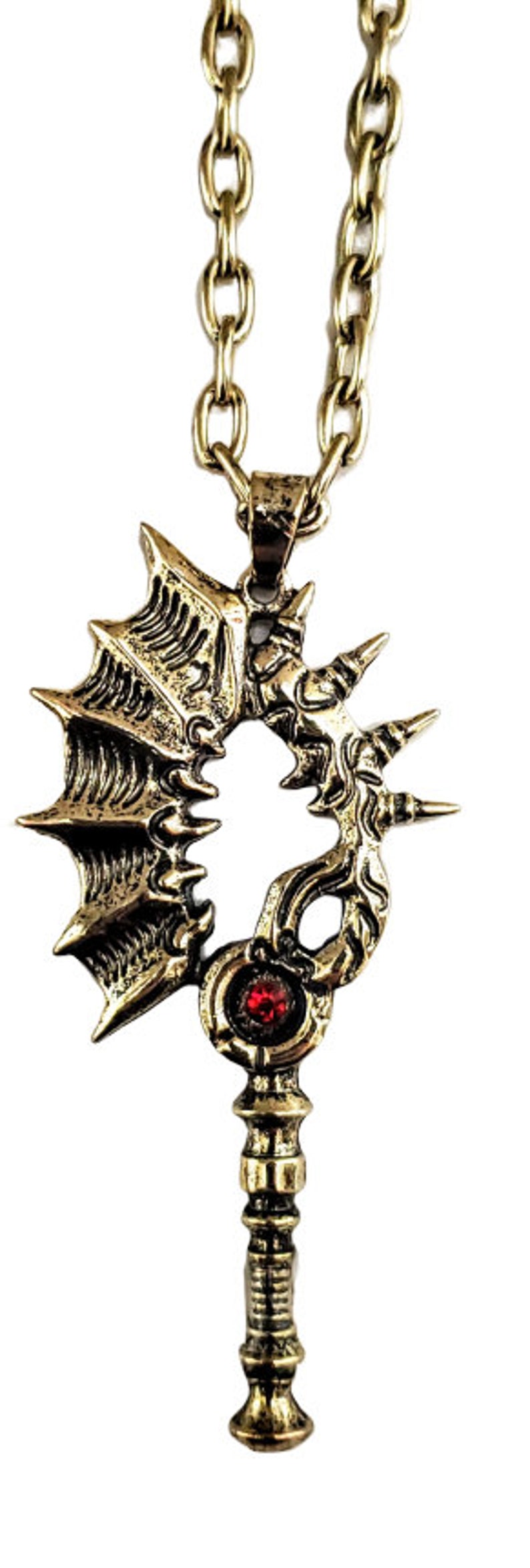May include: A bronze-colored pendant necklace featuring a stylized key design. The pendant has a detailed, ornate pattern with a red gemstone and is suspended from a matching chain.