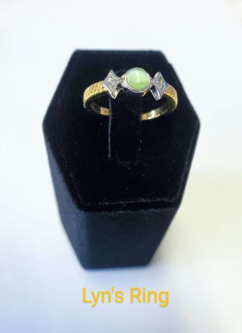 May include: A gold and silver ring with a light green gemstone center, flanked by two diamond-shaped accents. The ring is displayed on a black velvet ring box. The text "Lyn's Ring" is written in yellow.