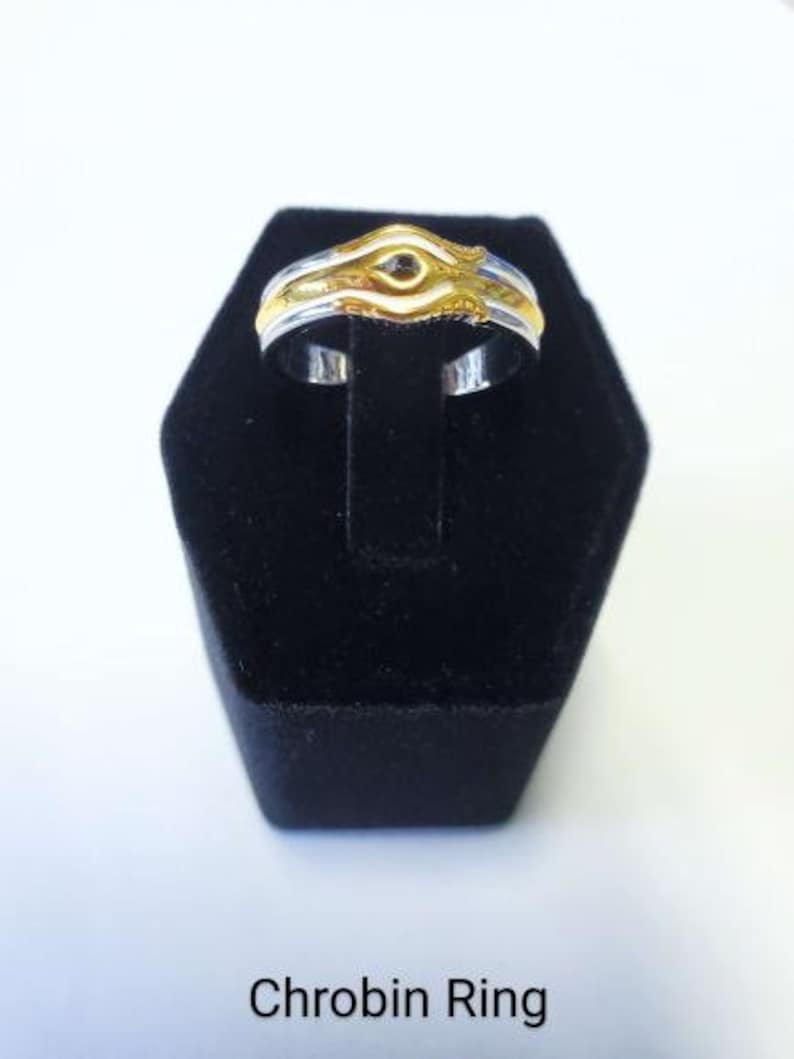 May include: A two-tone ring with a gold and silver design, displayed on a black stand. The ring features a gold centerpiece with a unique shape. The text "Chrobin Ring" is visible at the bottom of the image.