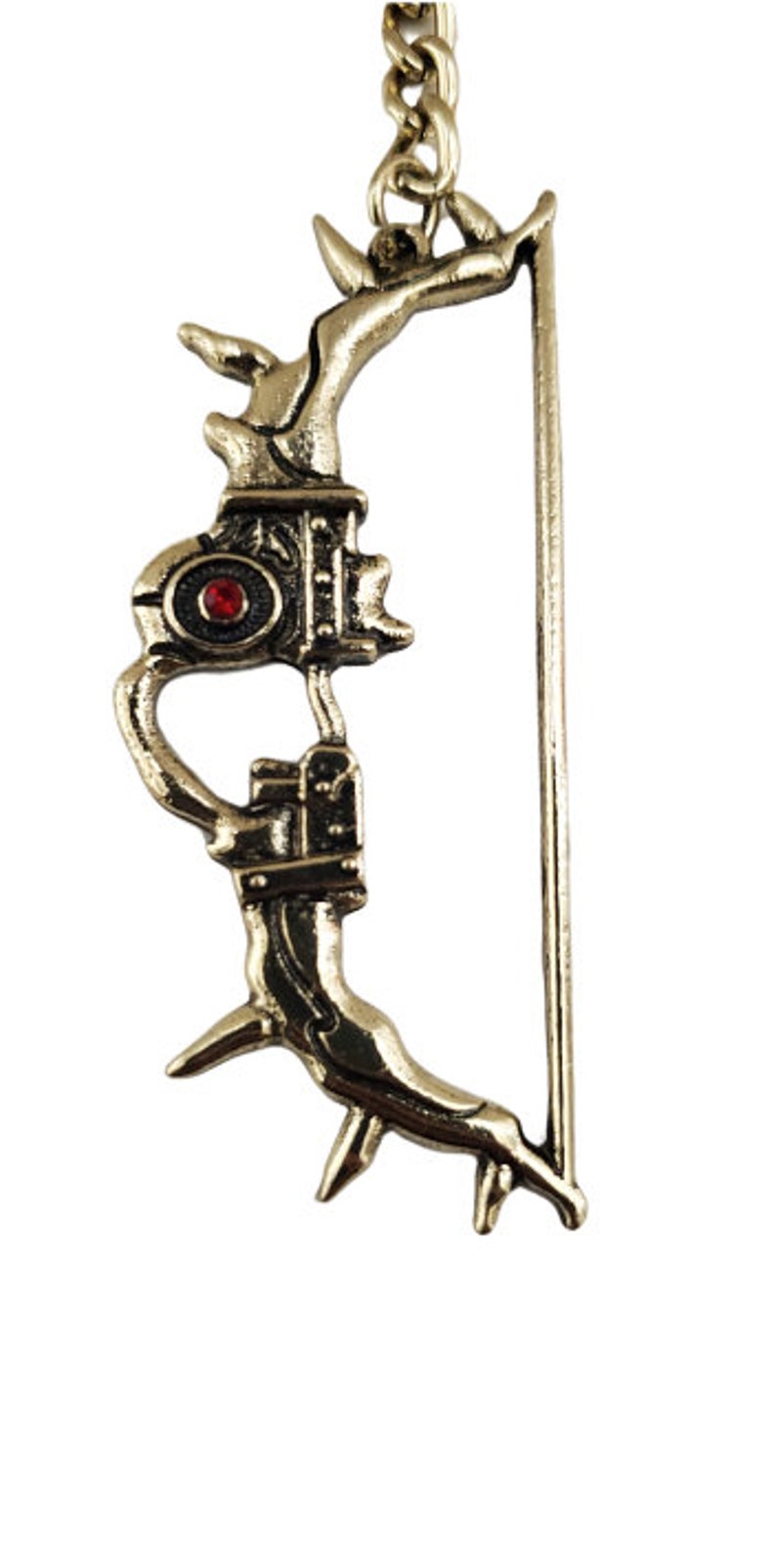 May include: A gold-colored bow and arrow keychain. The bow features intricate detailing and a red gem in the center. The arrow is a simple, straight design. The keychain is suspended from a small chain.