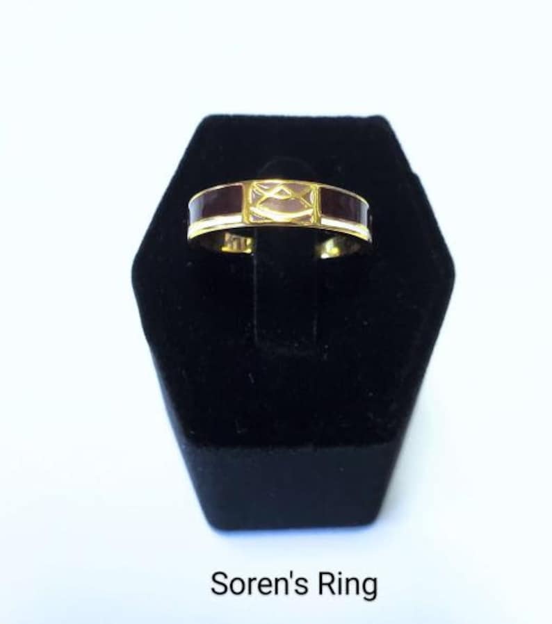 May include: A gold-colored ring with a rectangular design. The ring features dark burgundy enamel on the sides and a lighter, possibly mother-of-pearl, center section. The text "Soren's Ring" is at the bottom.