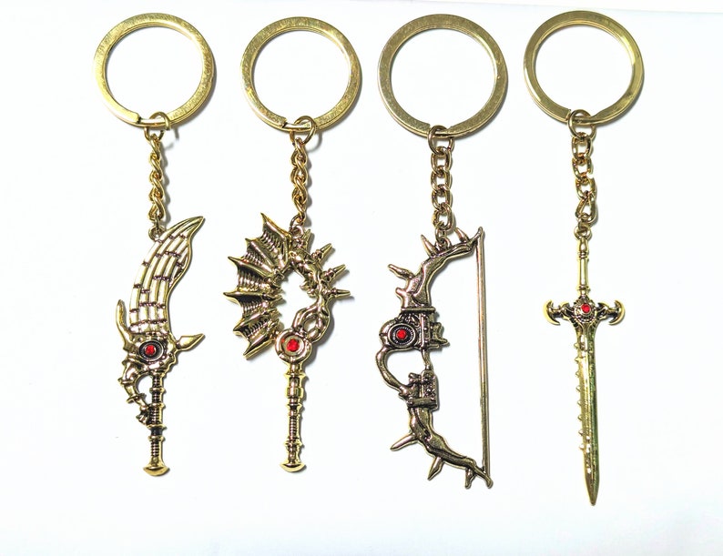 May include: Four gold-colored keychains with detailed fantasy weapon charms. The charms include a sword, a staff, a bow and arrow, and a sword. Each charm has a red gem accent and is attached to a gold chain and keyring.