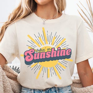May include: A white t-shirt with a graphic of a yellow sun with rays and the text "Walkin' on Sunshine" in pink and blue letters.