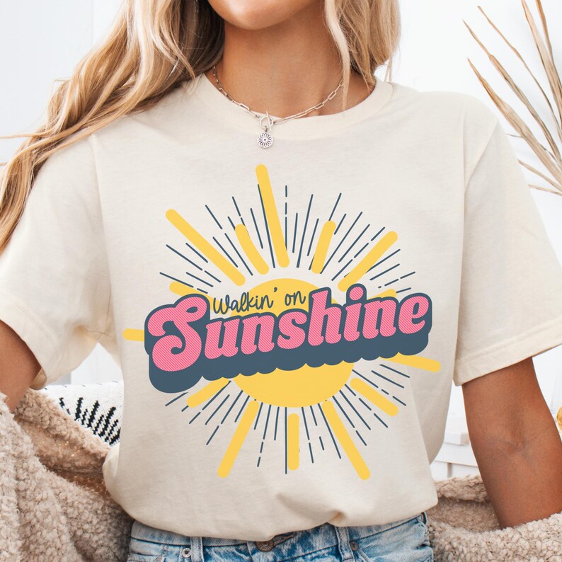 May include: A white t-shirt with a graphic of a yellow sun with rays and the text "Walkin' on Sunshine" in pink and blue letters.