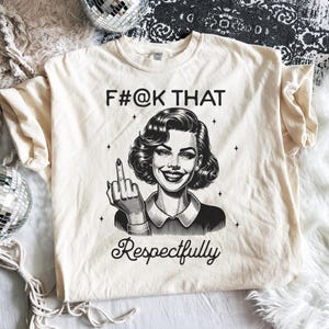 May include: A cream-coloured short-sleeve t-shirt featuring a black graphic illustration of a smiling person with wavy hair giving the middle finger. The text F#@K THAT is printed above the illustration and the word Respectfully is printed below.