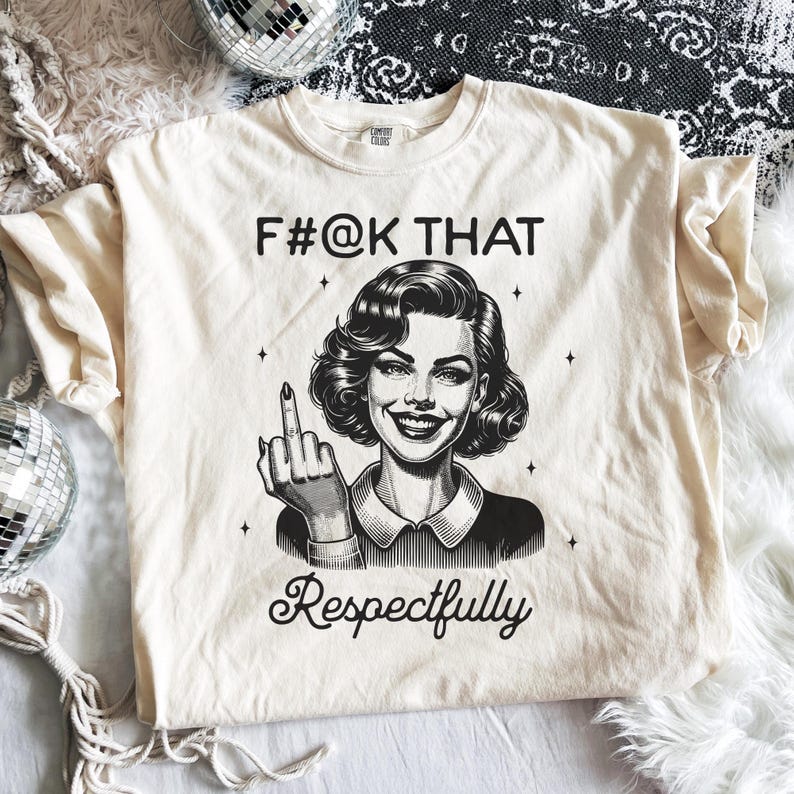 May include: A cream-coloured short-sleeve t-shirt featuring a black graphic illustration of a smiling person with wavy hair giving the middle finger. The text F#@K THAT is printed above the illustration and the word Respectfully is printed below.