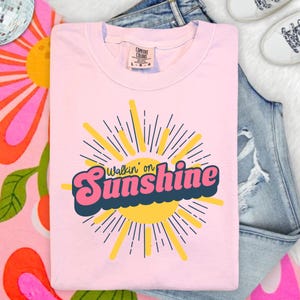 May include: A light pink short-sleeved t-shirt featuring a graphic of a yellow sun with radiating lines. The text "Walkin' on Sunshine" is printed across the center in a bold, pink, retro-style font with a dark blue outline and shadow.
