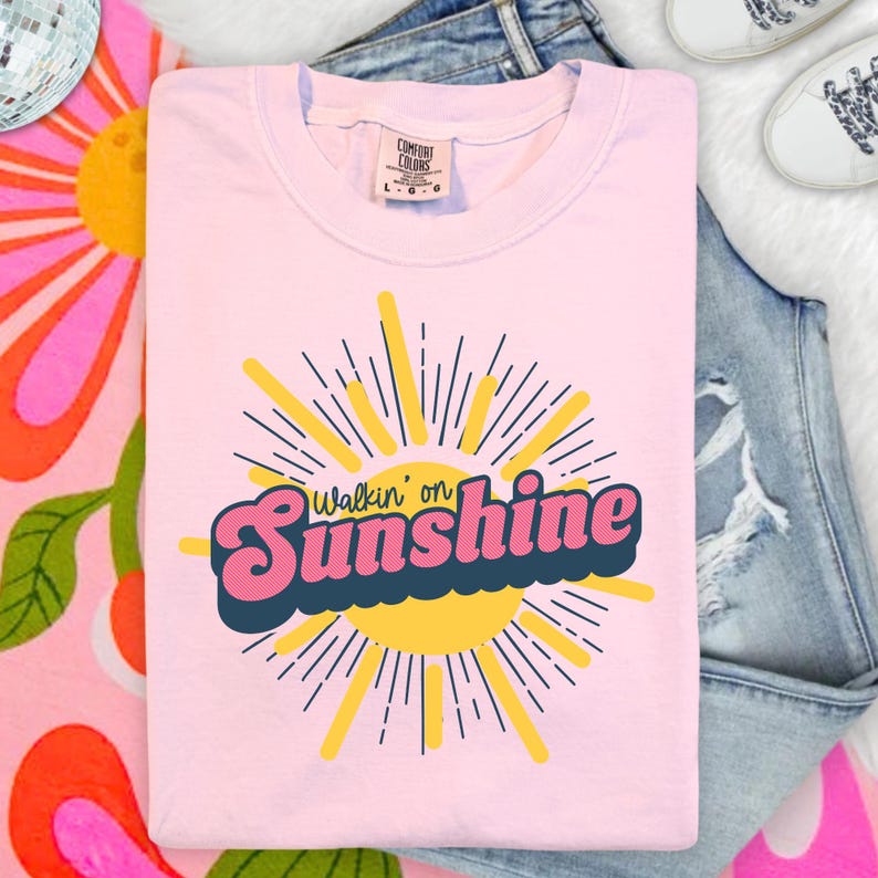 May include: A light pink short-sleeved t-shirt featuring a graphic of a yellow sun with radiating lines. The text "Walkin' on Sunshine" is printed across the center in a bold, pink, retro-style font with a dark blue outline and shadow.