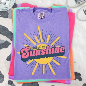 May include: A purple short-sleeve t-shirt featuring a yellow sun graphic with rays and the text Walkin' on Sunshine in pink and dark blue. The shirt has a Comfort Colors brand label at the inner neckline indicating a size medium.