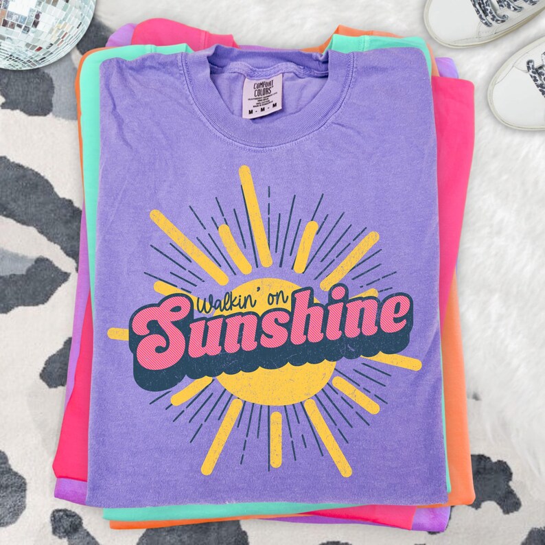 May include: A purple short-sleeve t-shirt featuring a yellow sun graphic with rays and the text Walkin' on Sunshine in pink and dark blue. The shirt has a Comfort Colors brand label at the inner neckline indicating a size medium.