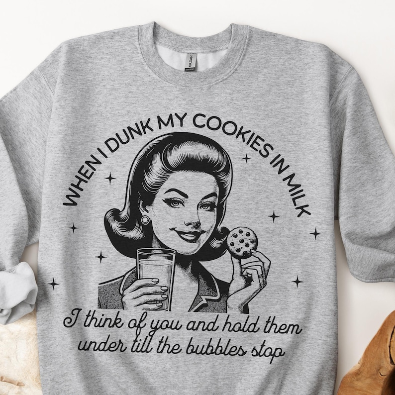 May include: A light grey crewneck sweatshirt featuring a black graphic illustration of a person holding a glass of milk and a chocolate chip cookie. The text reads: WHEN I DUNK MY COOKIES IN MILK I think of you and hold them under till the bubbles stop.