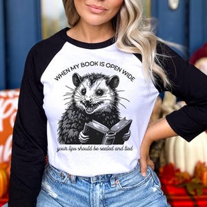 May include: A white and black baseball tee shirt with a graphic of a possum reading a book. The text on the shirt reads "When my book is open wide your lips should be sealed and tied."