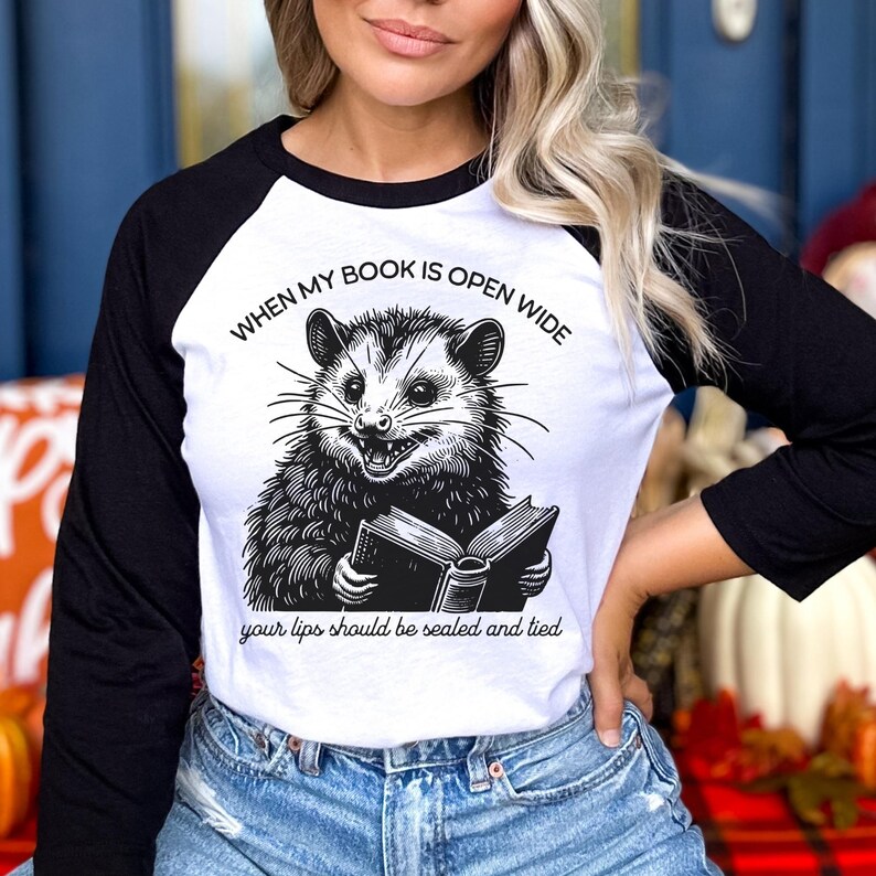 May include: A white and black baseball tee shirt with a graphic of a possum reading a book. The text on the shirt reads "When my book is open wide your lips should be sealed and tied."