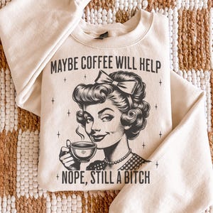 May include: A cream-coloured crewneck sweatshirt featuring a black graphic illustration of a person with a bow in their hair holding a steaming coffee cup. The text reads MAYBE COFFEE WILL HELP at the top and NOPE, STILL A BITCH at the bottom.