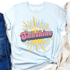 May include: Light blue t-shirt with a retro-style graphic of a sun with rays and the text "Walkin' on Sunshine".