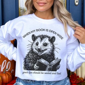 May include: White sweatshirt with a black and white illustration of an opossum reading a book. The text on the sweatshirt reads "When my book is open wide your lips should be sealed and tied."